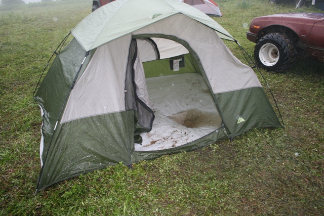 Green and tan tent on wet grass with part of a reddish vehicle visible.