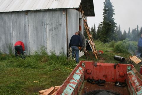 1990 Chevrolet Suburban - A group of people are working on a corrugated metal shed in a grassy, wooded area. One person is using a chainsaw near a ladder leaning aga…