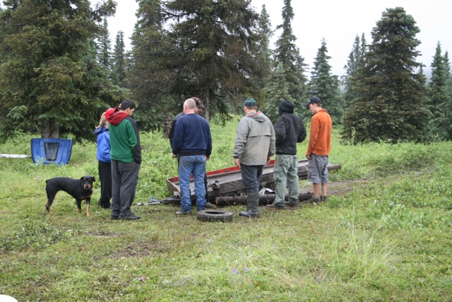 A group of people and a dog stand around a damaged trailer in a wooded area.