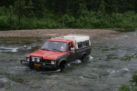 1990 Chevrolet Suburban - A red and grey pickup truck with a camper shell is driving through a shallow river. The driver is visible through the windshield, looking t…