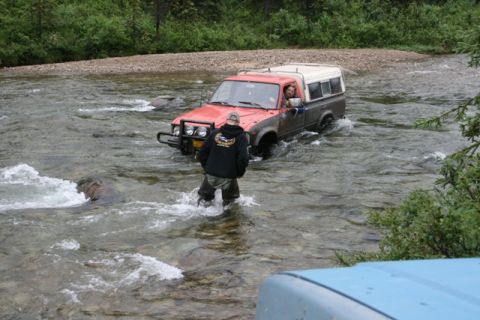 1990 Chevrolet Suburban - A red and white pickup truck with a camper shell is driving through a shallow river. A person in waders stands in the water, facing the tru…