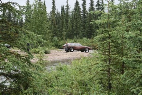 1990 Chevrolet Suburban - A brown station wagon with large tires is parked on a gravelly shore next to a stream, with a person standing beside it. The vehicle is sur…