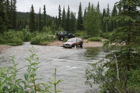 1990 Chevrolet Suburban - Two off-road vehicles are parked near a shallow river, with one SUV partially submerged in the water. The scene is set in a wooded area wit…