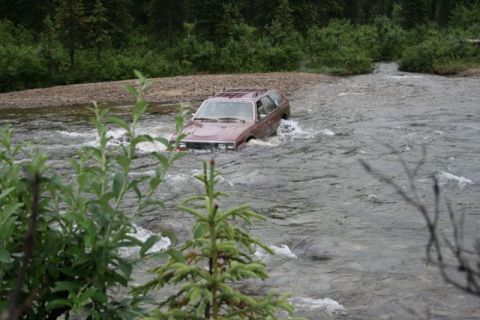 1990 Chevrolet Suburban - A maroon station wagon is partially submerged as it drives through a shallow, rocky river. Dense green trees and bushes line the river bank…