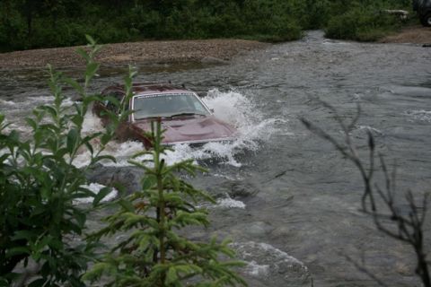 1990 Chevrolet Suburban - A maroon station wagon is driving through a shallow river, creating a large splash. The vehicle is partially obscured by foliage in the for…