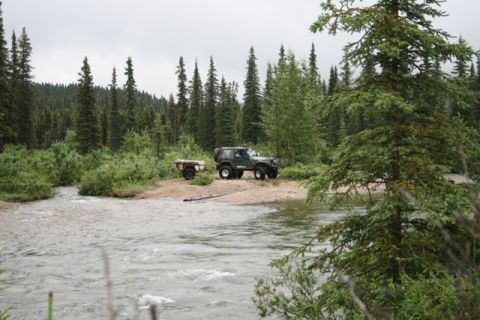 1990 Chevrolet Suburban - A dark green Jeep Wrangler is parked on a gravel shore next to a river, hitched to a small utility trailer. The vehicle is surrounded by de…