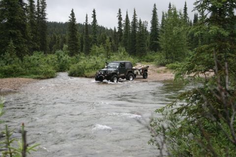 1990 Chevrolet Suburban - A dark green Jeep Wrangler is driving through a shallow river, pulling a small utility trailer loaded with wood. The vehicle is surrounded…