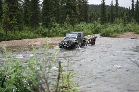 1990 Chevrolet Suburban - A dark green Jeep Wrangler is driving through a shallow river, pulling a small utility trailer loaded with wood. The vehicle is surrounded…