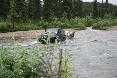 1990 Chevrolet Suburban - A dark green Jeep Wrangler is driving through a shallow river, with a wooded area and green bushes in the background. The Jeep has a rack o…