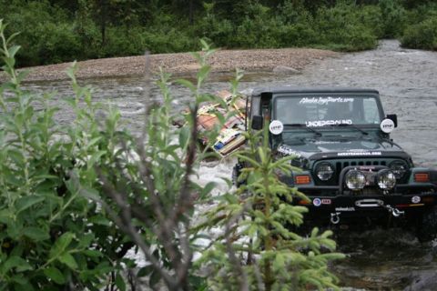 1990 Chevrolet Suburban - A dark green Jeep Wrangler is driving through a shallow river, carrying a large, strapped-down load on its roof rack. The Jeep has several…