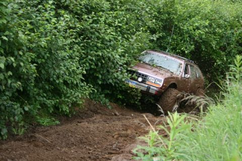 1990 Chevrolet Suburban - A brown AMC Eagle wagon is stuck in mud on a dirt track, surrounded by dense green foliage. The vehicle's tires are caked in mud, and it ap…