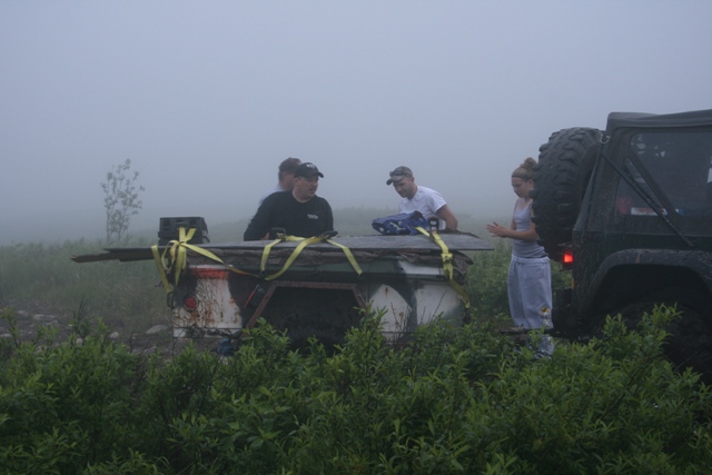 People around a trailer with lumber, a Jeep in the background, in a foggy outdoor setting.