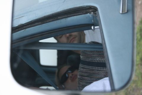 1990 Chevrolet Suburban - Reflection in a vehicle's side mirror shows two people sleeping inside the vehicle. A blue seatbelt is visible across the frame.