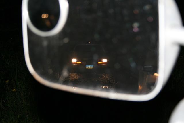 Rear view of a dark sedan with illuminated taillights and license plate, reflected in a side mirror at night.