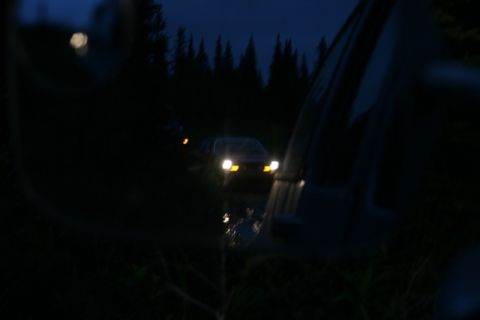 1990 Chevrolet Suburban - A dark sedan with its headlights on drives on a wet road at night, seen through a car's side mirror. The vehicle is surrounded by trees and…