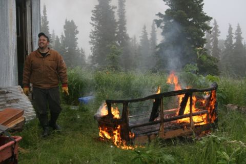 1990 Chevrolet Suburban - A man stands next to a couch that is on fire in a grassy, wooded area.