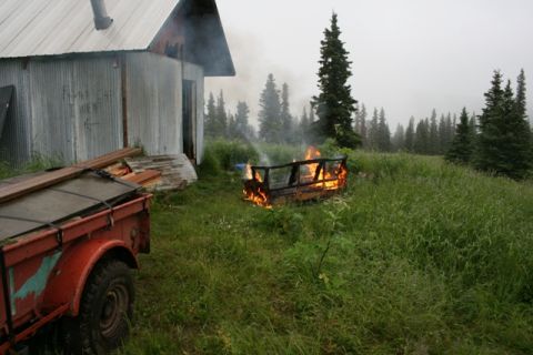 1990 Chevrolet Suburban - A red trailer with wood strapped to it is parked next to a corrugated metal building. A fire burns in a metal frame in the grassy yard.
