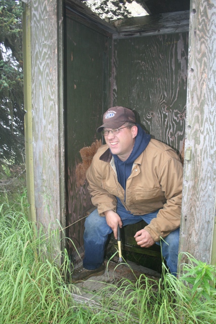 1990 Chevrolet Suburban - A man wearing a baseball cap and a brown jacket is sitting in the doorway of a weathered wooden structure, possibly an outhouse. He is hold…