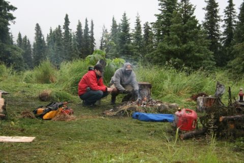 1990 Chevrolet Suburban - Two people are crouching around a campfire in a grassy, wooded area. A red gas can and camping gear are visible nearby.