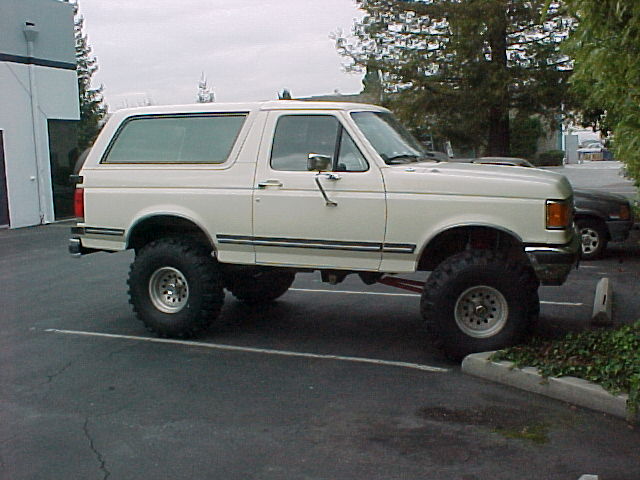 Lifted white SUV parked in a lot, side view. It has large tires and a raised suspension