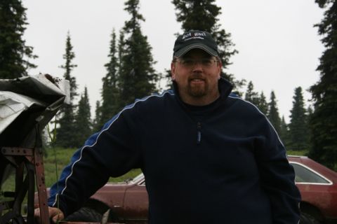 1990 Chevrolet Suburban - A man wearing a dark blue quarter-zip sweatshirt and a baseball cap stands in front of a red car with trees in the background. The man is l…
