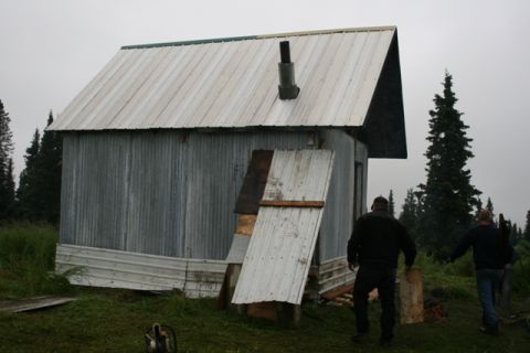 1990 Chevrolet Suburban - Two men walk away from a rustic, corrugated metal shed with a chimney, surrounded by trees on a cloudy day.