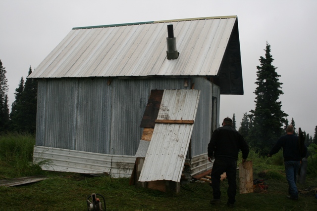 Two men walking away from a corrugated metal shed with a chimney in a wooded area.