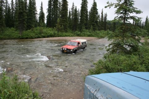 1990 Chevrolet Suburban - A red and brown pickup truck with a white camper shell is driving through a shallow river surrounded by dense evergreen trees.