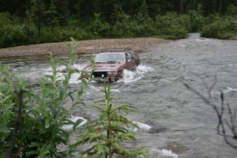 1990 Chevrolet Suburban - A maroon station wagon drives through a shallow river with trees and brush lining the banks.