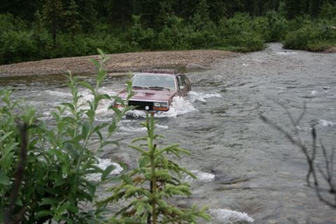 1990 Chevrolet Suburban - A maroon AMC Eagle wagon drives through a shallow, rocky river. The vehicle is partially submerged, with water splashing around its tires a…