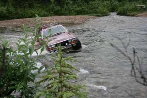 1990 Chevrolet Suburban - A maroon SUV is driving through a shallow river with trees and brush on the banks.