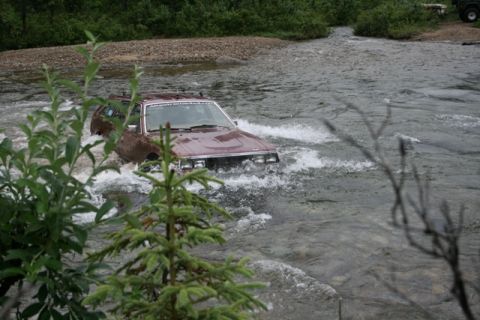 1990 Chevrolet Suburban - A maroon station wagon is driving through a shallow river, creating splashes. The vehicle is partially obscured by foliage in the foregroun…