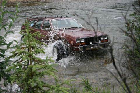 1990 Chevrolet Suburban - A maroon AMC Eagle wagon drives through a shallow river, kicking up a large spray of water. The vehicle has off-road tires and a roof rack.
