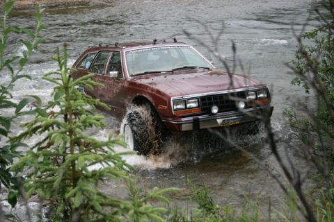 1990 Chevrolet Suburban - A maroon AMC Eagle wagon is driving through a shallow river, kicking up water. The vehicle has large off-road tires and a roof rack.