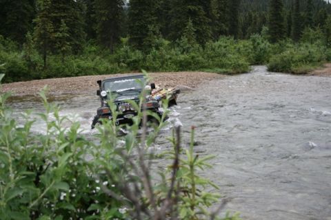 1990 Chevrolet Suburban - A dark green Jeep Wrangler is driving through a shallow, rocky river with trees and brush on the banks.