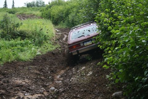 1990 Chevrolet Suburban - A maroon station wagon is driving down a steep, muddy, off-road trail, with its tires kicking up mud. The vehicle is partially obscured by…