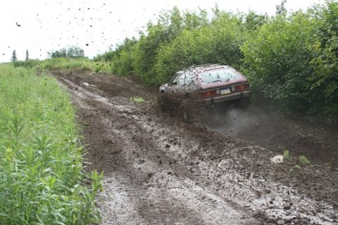 1990 Chevrolet Suburban - A brown station wagon is driving up a steep, muddy hill, kicking up a lot of mud and dirt. The vehicle is covered in mud, and the surroundi…