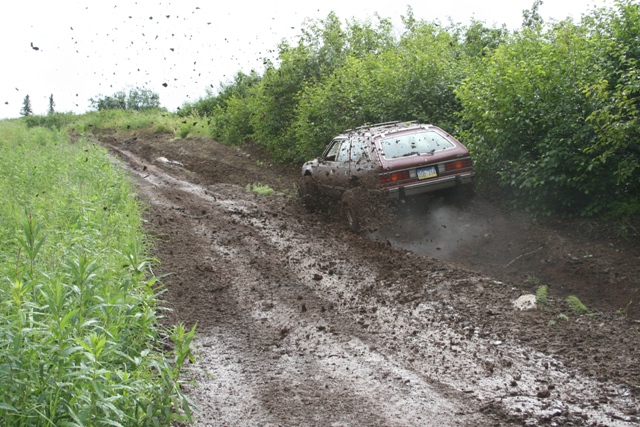 Brown station wagon driving up a muddy hill, covered in mud.