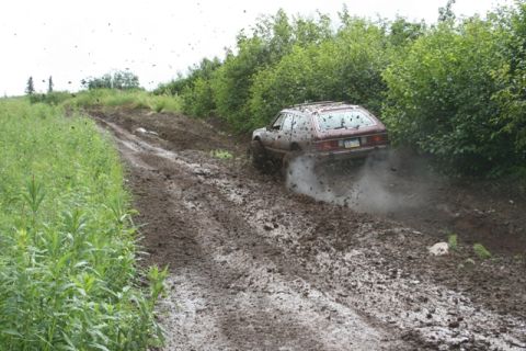 1990 Chevrolet Suburban - A maroon station wagon navigates a steep, muddy off-road trail, kicking up dirt and mud as it goes. The vehicle is surrounded by lush green…