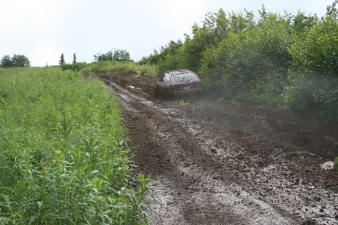 1990 Chevrolet Suburban - A red station wagon is stuck in deep mud on a dirt road, surrounded by tall green grass and trees.