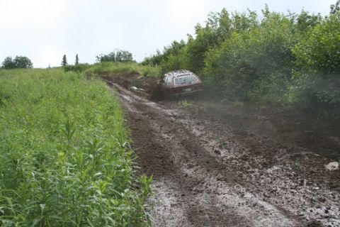 1990 Chevrolet Suburban - A red station wagon is stuck in thick mud on a steep, unpaved road. The vehicle is tilted and appears to be struggling to gain traction.