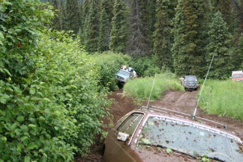 1990 Chevrolet Suburban - A brown station wagon, heavily covered in mud, is stuck on a muddy track surrounded by dense green foliage and tall evergreen trees. Severa…