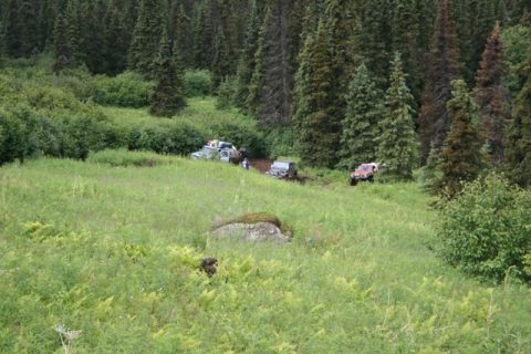 1990 Chevrolet Suburban - Several off-road vehicles are navigating a grassy, wooded area with some muddy patches. One red SUV is visible on the right side of the fra…