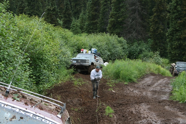 Light blue Chevrolet Suburban stuck in mud on a steep, wooded trail with a man holding a rope.