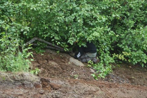 1990 Chevrolet Suburban - A person in a black hoodie and waders is crouched down behind dense green foliage, partially obscured by a metal structure. The scene appea…