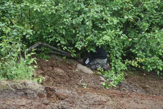 Person in black hoodie and waders crouching behind foliage and metal structure on a muddy slope.
