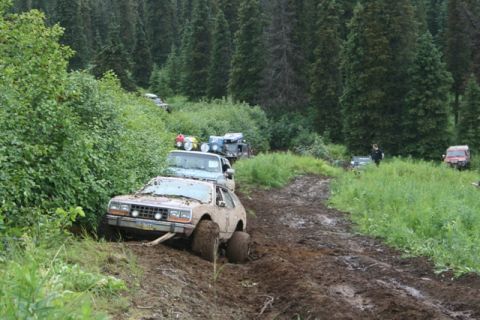 1990 Chevrolet Suburban - Several off-road vehicles navigate a muddy, rutted trail through a dense forest. The lead vehicle, a light-colored SUV, is partially submer…