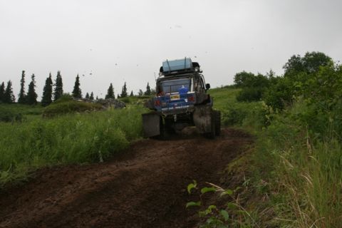 1990 Chevrolet Suburban - A modified blue Chevrolet Suburban drives up a muddy trail with a blue cooler on its roof rack. The vehicle has large tires and mud flaps,…
