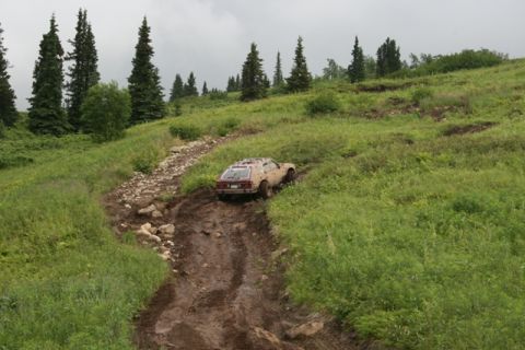 1990 Chevrolet Suburban - A tan, dusty station wagon is navigating a steep, muddy, and rocky off-road trail on a grassy hillside under an overcast sky. The vehicle a…