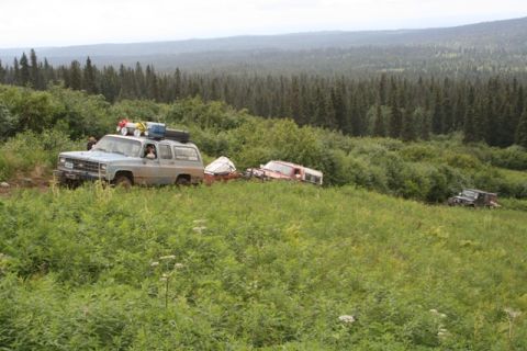 1990 Chevrolet Suburban - A light blue Chevrolet Suburban tows a trailer with a red vehicle on it, driving through tall green grass and dense forest. Another dark co…